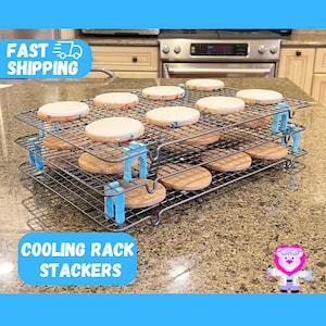 May include: A three-tiered cooling rack with blue stackers, holding iced cookies. The cookies have white frosting and colorful sprinkles. The rack is made of metal wire. The text "COOLING RACK STACKERS" is visible.