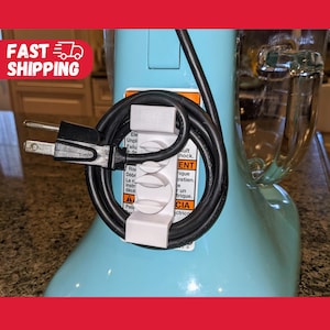 May include: A white cord organizer with a black electrical cord wrapped around it. The organizer is attached to a light blue kitchen appliance with a black warning label that reads: "Ele Unpl Failu In de SULT HOCK. ENT Risi rique Debr tretien. Len le instr dece A CIA Peli ctrico B".