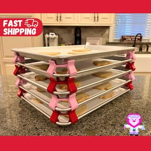 May include: Four baking sheets are stacked on top of each other, secured with pink and red clips. The baking sheets are made of metal and have a silver finish. The clips are designed to hold the baking sheets together and prevent them from sliding.