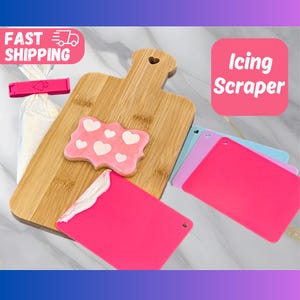 May include: A set of four pink, purple, and blue silicone icing scrapers on a wooden cutting board. The scrapers have a heart-shaped cutout in the top corner. A pink cookie with white hearts is on the cutting board. The text "Icing Scraper" is in a pink box.