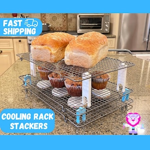 May include: Two silver cooling racks stacked on top of each other with white clips holding them together. The top rack has two loaves of white bread on it. The bottom rack has six muffins on it. The racks are on a gray countertop.