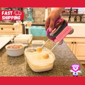 May include: A pink and gray handheld kitchen tool labeled POP CLIPS with two white prongs holding round cake pops. The tool is positioned over a clear plastic container filled with white icing, while a bowl of colorful sprinkles sits nearby.