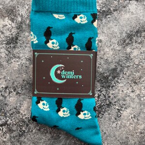 May include: A pair of teal blue socks with a pattern of white skulls and black crows. The socks have a tag that says "demi winters" with a crescent moon and stars.