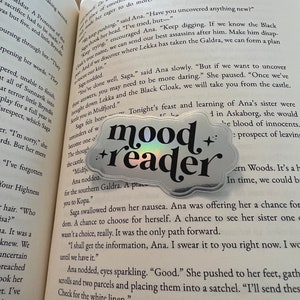 Könnte beinhalten: Nahaufnahme eines Buches mit einem Aufkleber, auf dem "mood reader" steht. Das Buch ist auf einer Seite geöffnet, auf der Text über eine Figur namens Saga und einen Ort namens Southern Galdra steht.