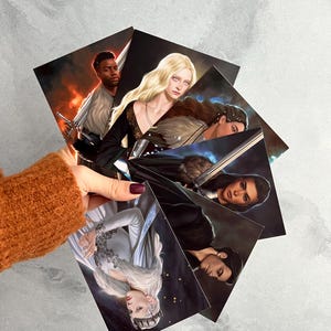 Könnte beinhalten: Ein Set aus sechs illustrierten Karten mit Charakteren aus einem Fantasy-Roman. Die Karten sind auf dickem Karton gedruckt und zeigen leuchtende Farben und komplizierte Details. Jede Karte zeigt einen anderen Charakter, darunter eine Frau mit langen blonden Haaren, ein Mann mit dunklen Haaren und eine Frau mit dunklen Haaren und einem Schwert.