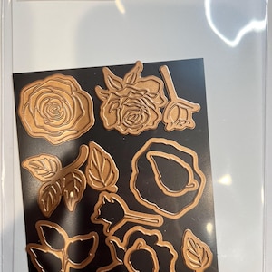 May include: A set of Spellbinder-Glimmer Edge Roses die cuts in a clear plastic package. The die cuts are in various shapes of roses and leaves, made of a copper-colored metal. The shapes are arranged on a black background.
