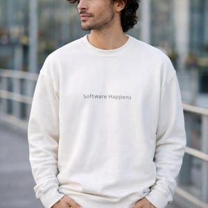 Peut inclure: Sweat-shirt crème avec l'inscription "Software Happens" en lettres simples. Le sweat-shirt est de style col rond et est porté avec un jean bleu. La personne se tient à l'extérieur.