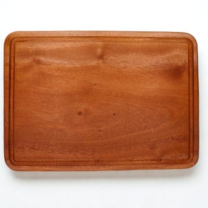 Puede incluir: Tabla de cortar rectangular de madera con acabado marrón rojizo. La tabla tiene un borde empotrado alrededor de los bordes. La veta de la madera es visible y la tabla está sobre un fondo blanco. Ideal para la preparación de alimentos.