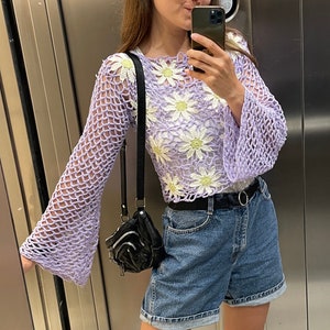 Pode incluir: Uma mulher vestindo um top de crochê roxo claro com flores brancas de margarida, shorts jeans azuis e sandálias pretas. Ela está em um elevador e tirando uma selfie com um smartphone preto.