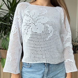 Puede incluir: Un suéter de crochet azul claro de tejido abierto, con mangas largas y cuello barco. El suéter se combina con pantalones cortos vaqueros azules claros. El fondo incluye plantas verdes y muebles de madera.