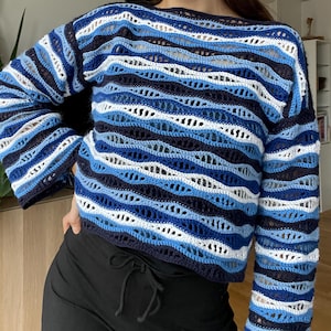 May include: A blue and white crochet sweater with long sleeves. The sweater has a striped pattern in shades of blue and white. The neckline is a simple crew neck. The sleeves are long and the fit is relaxed.