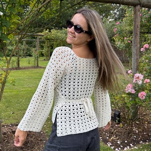 May include: A cream-colored long-sleeve crochet top featuring an open-knit pattern, a square neckline, and a tie-back closure at the waist, paired with dark gray denim shorts. The top has flared sleeves and a side-slit hemline design.