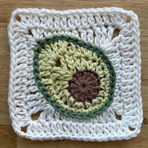Puede incluir: Un cuadrado de crochet blanco con un diseño de aguacate verde, amarillo y marrón en el centro.