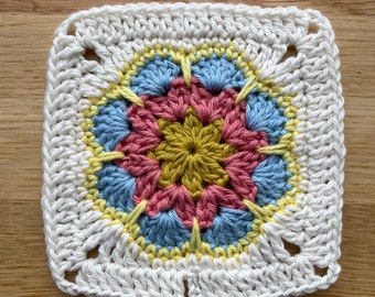 May include: A white crochet square with a colorful flower design in the center. The flower has yellow, pink, and blue petals.