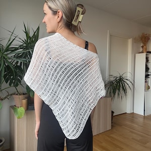 Może przedstawiać: Biała, ażurowa chusta z geometrycznym wzorem. Chusta jest zarzucona na ramię. Osoba ma na sobie czarne spodnie i czarny top. Złota spinka do włosów jest widoczna we włosach.