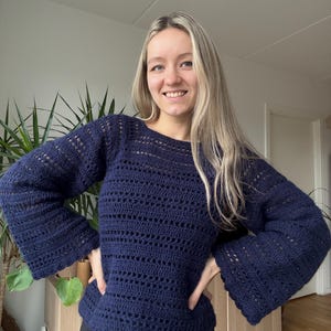 May include: A navy blue crochet sweater with long, flared sleeves. The sweater features a boat neck and an openwork design. The garment is handmade and appears to be a comfortable, stylish piece of clothing.