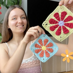 May include: Two crocheted granny squares with a retro daisy design. One square is green with a red flower and the other is blue with an orange flower.