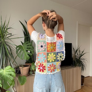 Op de afbeelding: Een kleurrijk gehaakt vest met een bloemenpatroon. Het vest is gemaakt van vierkanten met verschillende gekleurde bloemen. Het vest wordt gedragen over een wit T-shirt en een blauwe jeans.