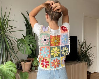 Può includere: Un gilet di uncinetto colorato con un motivo floreale. Il gilet è composto da quadrati con fiori di diversi colori. Il gilet è indossato sopra una maglietta bianca e jeans blu.