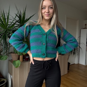Può includere: Un cardigan all'uncinetto a righe verdi e blu con scollo a V e chiusura con bottoni. Il maglione a maniche lunghe è abbinato a pantaloni neri. Il cardigan è fatto a mano e presenta un design unico e strutturato.