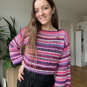 Könnte beinhalten: Ein gestreiftes Häkeltop mit lockerer Passform und langen Ärmeln. Das Top ist aus verschiedenen rosa, lila und weißen Garnen gefertigt.
