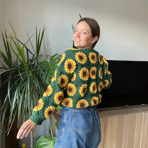 Könnte beinhalten: Eine grüne Häkeljacke mit Sonnenblumenmuster. Die Jacke ist kurz und hat lange Ärmel. Das Model trägt eine Jeans-Shorts.