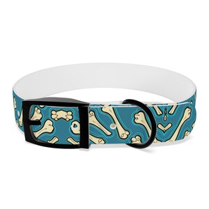 Op de afbeelding: Een witte hondenriem met een blauwe achtergrond en een patroon van witte botten. De riem heeft een zwarte gesp en een zilveren D-ring.