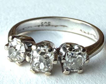 Puede incluir: Un anillo de plata con tres piedras preciosas redondas y transparentes. La banda del anillo es lisa y tiene un sello distintivo. Las piedras están engastadas en un engaste de garras clásico.