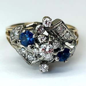 Könnte beinhalten: Ein Vintage-Goldring mit einem Cluster aus Edelsteinen. Der Ring verfügt über eine zentrale Anordnung aus runden und marquiseförmigen Diamanten, die mit leuchtenden blauen Saphiren durchsetzt sind. Die Fassung ist eine Mischung aus Gold und silberfarbenem Metall.