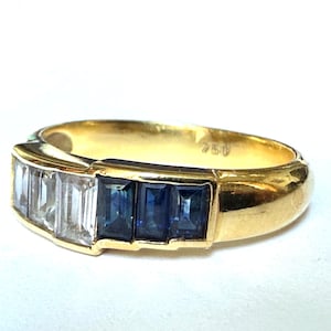 May include: A gold ring with a band featuring a row of rectangular clear stones, followed by three rectangular blue stones. The ring is marked with "750" on the inside.