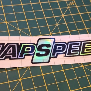 May include: A black and white sticker with the text "JAPSPEED" in a rainbow iridescent font.