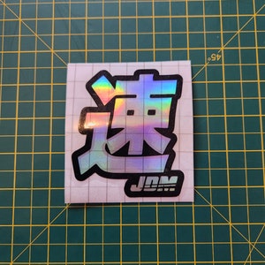 May include: A holographic sticker with the Japanese word "Drift" in black and white with a black "JDM" logo below it.