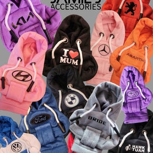 May include: A collection of miniature hoodie-style gear shifter covers in various colours including purple, pink, black, orange, blue, and grey. Each features white drawstrings and distinct logos or text like Kia, I Love Mum, Mercedes, Abarth, Hyundai, BMW, Ford, Bride, Subaru, VW, and Hawk Tuah.