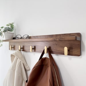 May include: A wooden wall-mounted shelf with brass hooks. The shelf is made of dark wood and has a small ledge. A pair of eyeglasses and a small potted plant sit on the shelf. Two coats and a bag hang from the hooks.