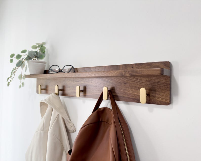 May include: A wooden wall-mounted shelf with brass hooks. The shelf is made of dark wood and has a small ledge. A pair of eyeglasses and a small potted plant sit on the shelf. Two coats and a bag hang from the hooks.