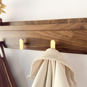May include: A wooden wall shelf with a dark brown finish and three gold-colored hooks. A light-colored jacket hangs from one hook, and a brown purse hangs from another. A small brown glass bottle with dried leaves sits on the shelf.