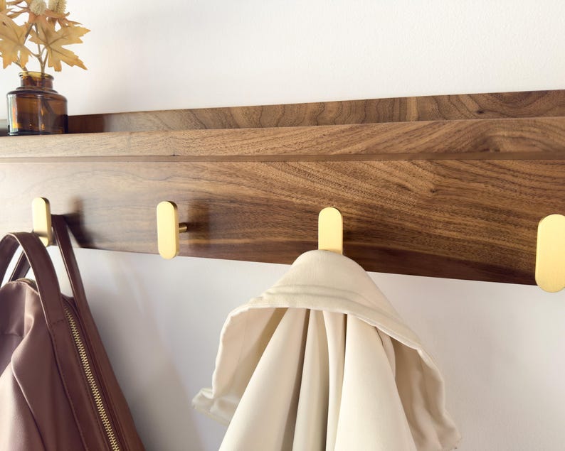 May include: A wooden wall shelf with a dark brown finish and three gold-colored hooks. A light-colored jacket hangs from one hook, and a brown purse hangs from another. A small brown glass bottle with dried leaves sits on the shelf.