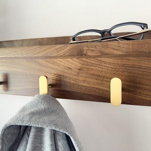 May include: A wooden wall-mounted shelf with a dark brown finish, featuring three gold-colored hooks. A pair of black-framed glasses rests on the shelf, and a gray hooded garment hangs from one of the hooks. The shelf is mounted on a white wall.