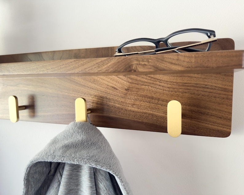 May include: A wooden wall-mounted shelf with a dark brown finish, featuring three gold-colored hooks. A pair of black-framed glasses rests on the shelf, and a gray hooded garment hangs from one of the hooks. The shelf is mounted on a white wall.