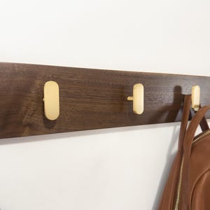 May include: A wooden wall-mounted coat rack with five brass hooks. The dark brown wood contrasts with the gold-colored hooks. A brown leather backpack hangs from one of the hooks. The rack is mounted on a white wall.