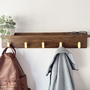 May include: A wooden wall shelf with four brass hooks. A brown leather backpack and a gray jacket hang from the hooks. A small potted plant and eyeglasses rest on the shelf. The shelf is mounted on a white wall.