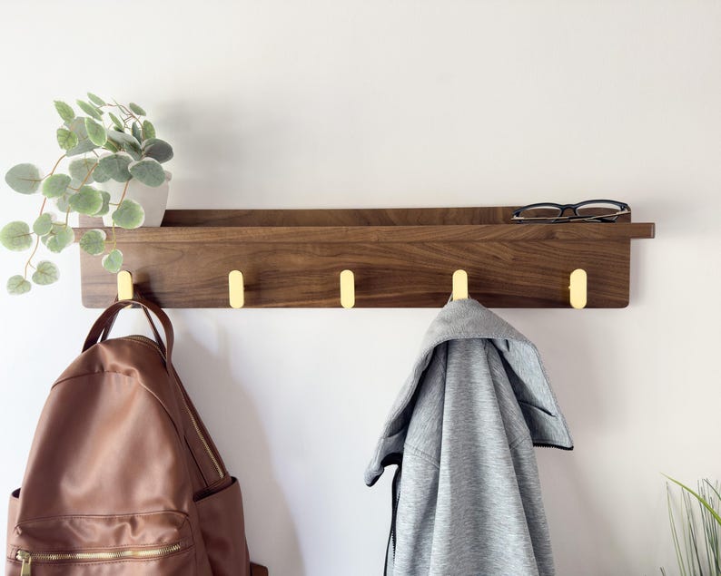 May include: A wooden wall shelf with four brass hooks. A brown leather backpack and a gray jacket hang from the hooks. A small potted plant and eyeglasses rest on the shelf. The shelf is mounted on a white wall.