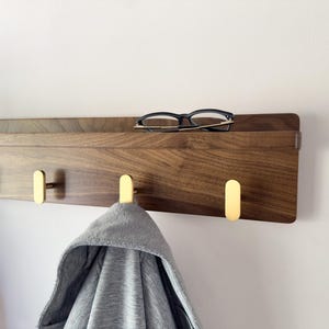 May include: A wooden wall-mounted shelf with three brass hooks. The shelf is made of dark wood and has a small ledge. A pair of black-framed glasses rests on the shelf. A gray hooded sweatshirt hangs from one of the hooks.
