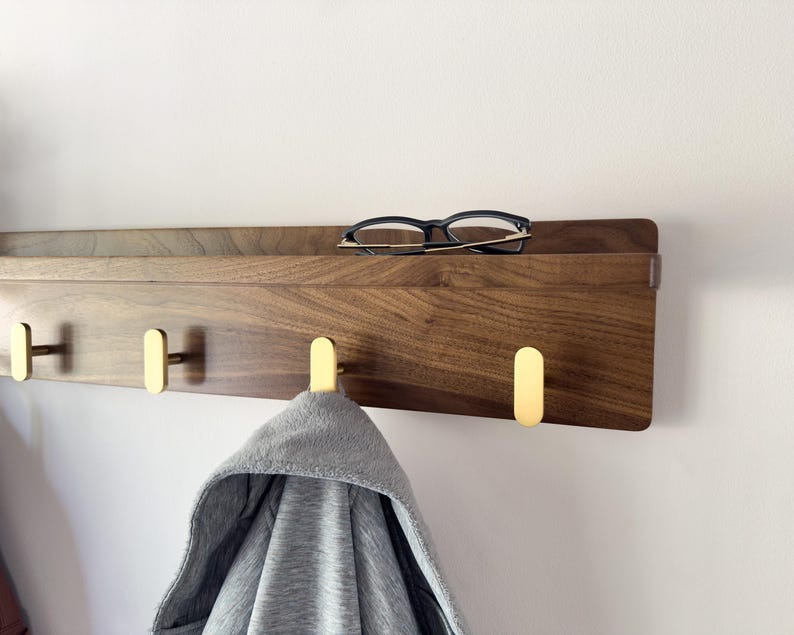 May include: A wooden wall-mounted shelf with three brass hooks. The shelf is made of dark wood and has a small ledge. A pair of black-framed glasses rests on the shelf. A gray hooded sweatshirt hangs from one of the hooks.