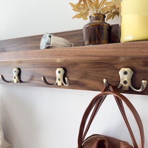 May include: A wooden wall-mounted shelf with three hooks and a small shelf. The shelf is made of dark wood and has a natural finish. The hooks are made of metal and have a brass finish. A brown leather bag hangs from one of the hooks.