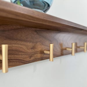 May include: A wooden shelf with five gold metal hooks. The shelf is made of walnut wood and has a natural finish. The hooks are simple and modern in design.