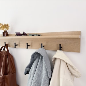May include: A wooden wall-mounted coat rack with a shelf and five hooks. The rack is made of light wood and has black metal hooks. There are three items hanging on the hooks, including a brown backpack, a gray jacket, and a white jacket.