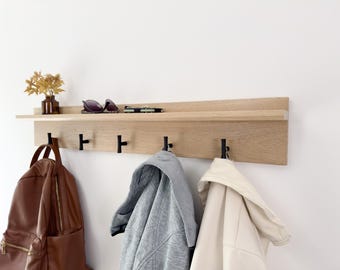 Puede incluir: Un perchero de pared de madera con un estante y cinco ganchos. El perchero está hecho de madera clara y tiene ganchos de metal negro. Hay tres artículos colgados en los ganchos, incluyendo una mochila marrón, una chaqueta gris y una chaqueta blanca.