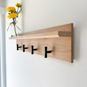 May include: A wooden wall-mounted shelf with five black hooks. The shelf is made of light-colored wood and has a small ledge. A glass vase with yellow flowers sits on the shelf.