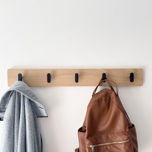 May include: A wooden coat rack with four black hooks mounted on a white wall. A gray hooded jacket hangs on one hook, and a brown leather backpack hangs on another. The rack is made of light-colored wood.
